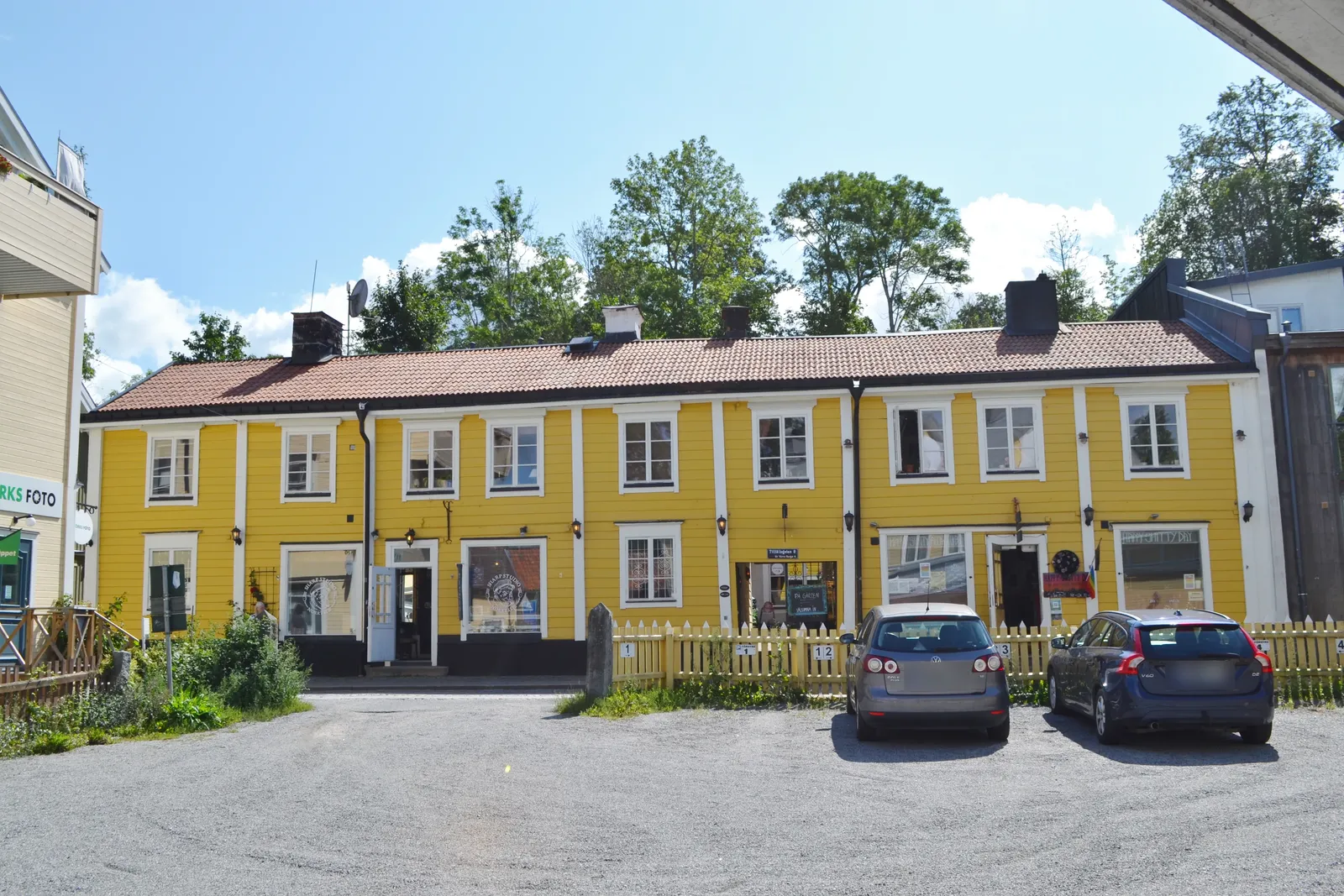 Gul trähusfasad i två våningar med butiksfönster i bottenvåningen och två bilar parkerade på en grusplan framför, med bakomliggande träd.