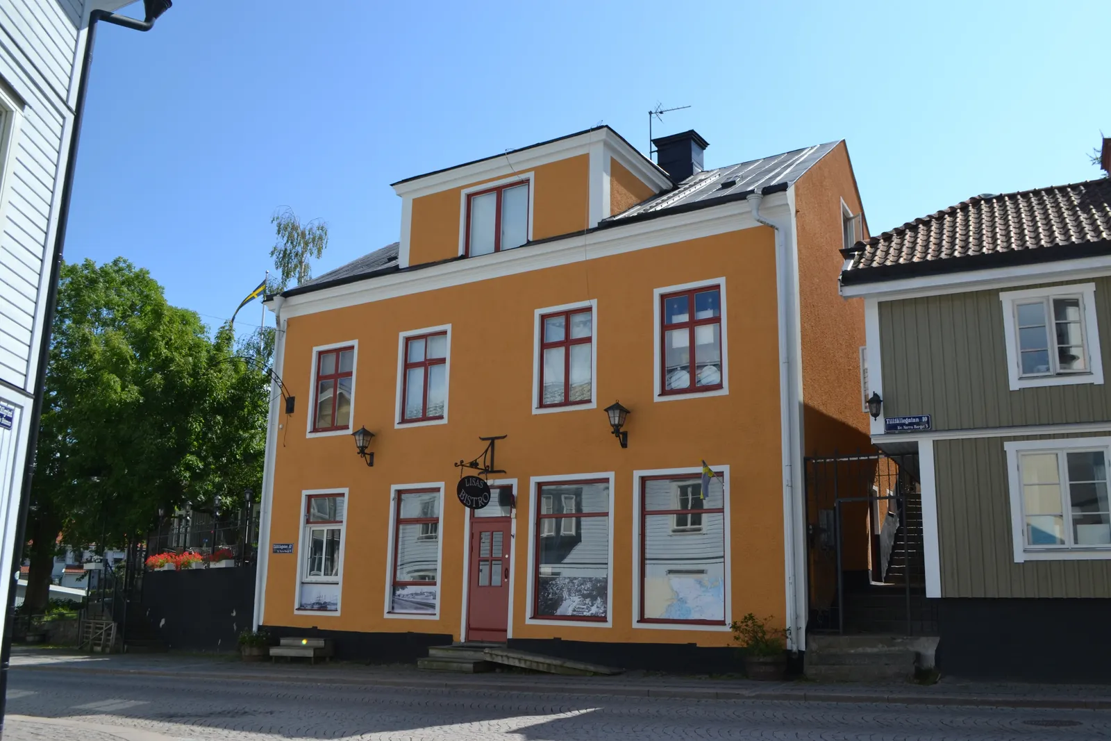 Orange putsad byggnad i två våningar med röda fönsterbågar och en skylt vid ingången, sedd från en kullerstensgata.