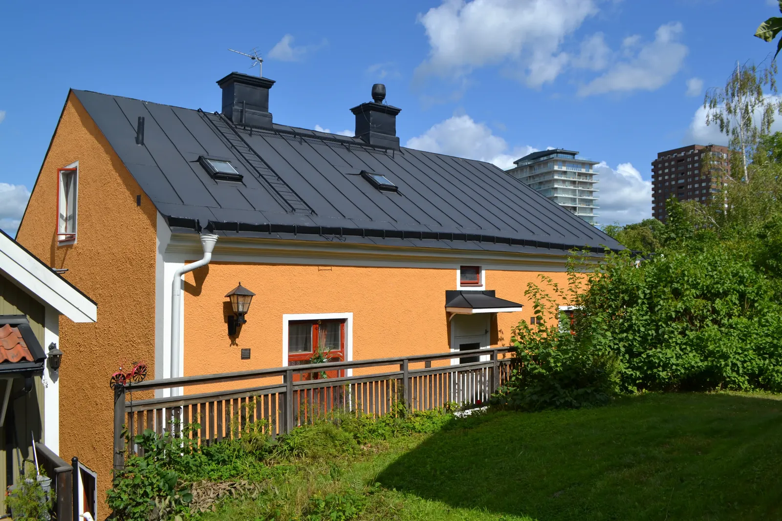 Orange putsad byggnad med svart plåttak, ett staket och en gräsyta framför, med höghus i bakgrunden.