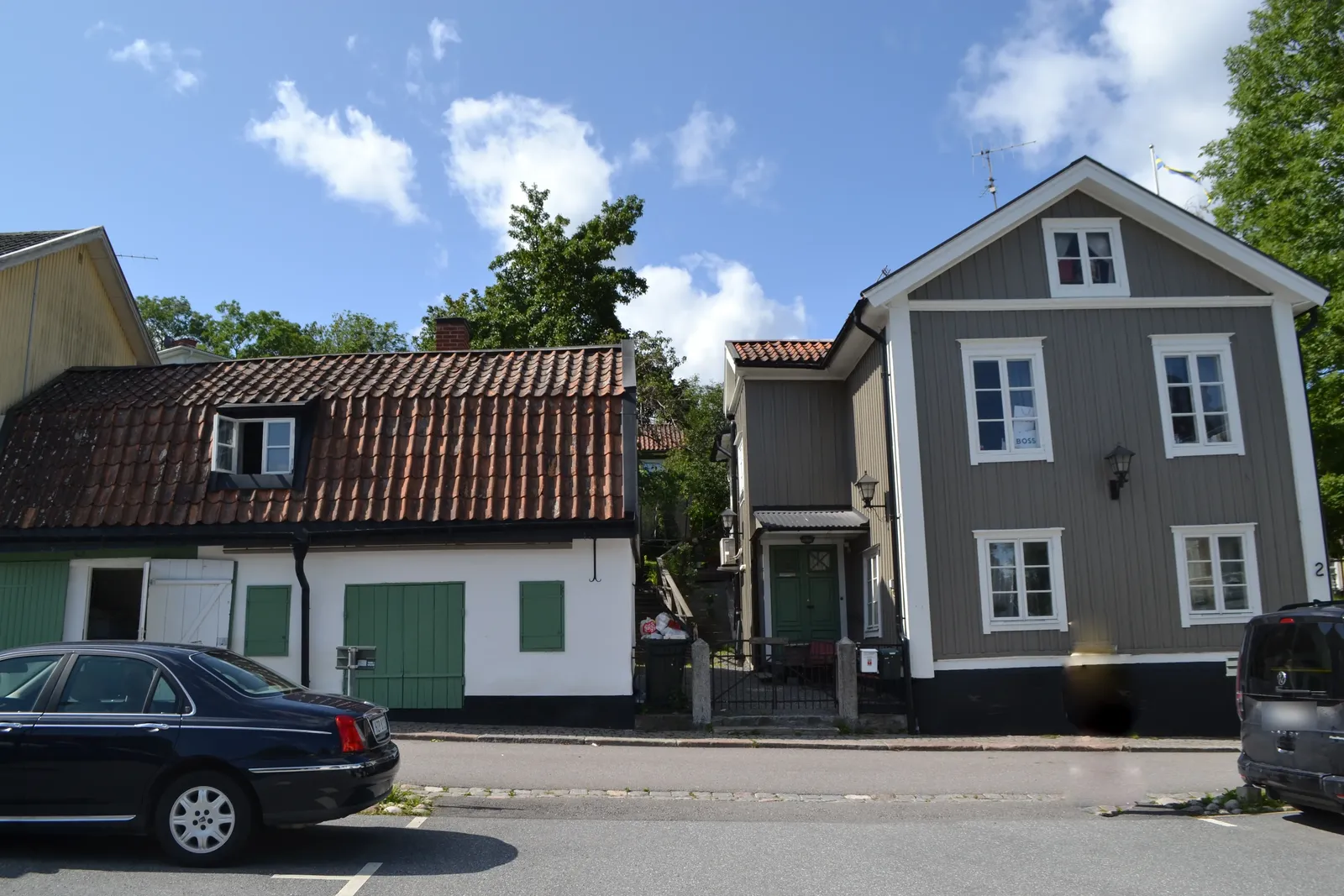Låg byggnad med tegeltak och vit fasad till vänster och en grå tvåvåningsbyggnad i trä till höger, med gata och parkerade bilar framför.